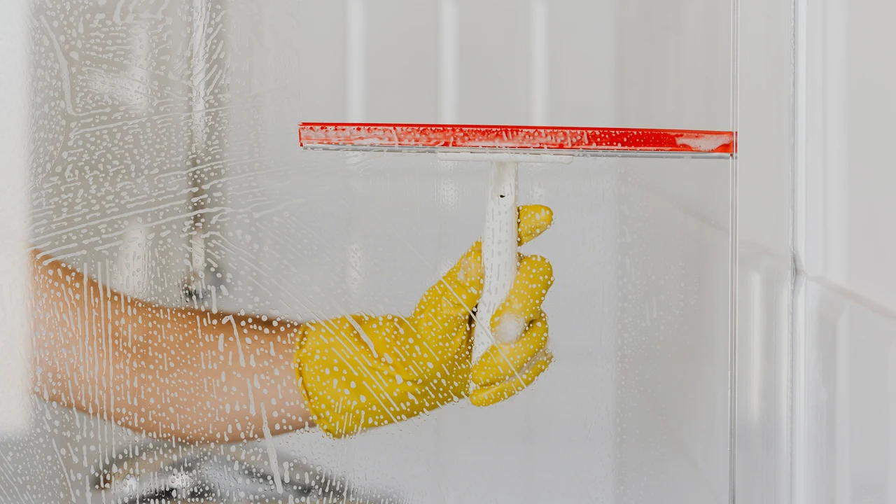 A gloved hand drawing a squeegee across a wet glass shower screen
